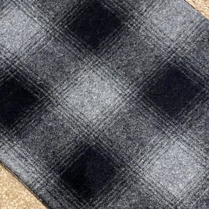 May include: Close-up of a fabric with a plaid pattern in shades of gray and black. The pattern features large, square blocks of dark gray and black, intersected by lighter gray lines. The fabric has a textured appearance.
