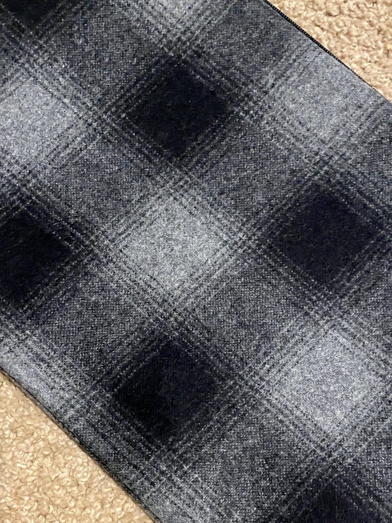 May include: Close-up of a fabric with a plaid pattern in shades of gray and black. The pattern features large, square blocks of dark gray and black, intersected by lighter gray lines. The fabric has a textured appearance.