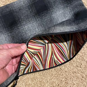 May include: A gray plaid zippered pouch with a black zipper. The interior lining features a colorful striped pattern in red, yellow, gray, and brown. The pouch is open, revealing the lining.