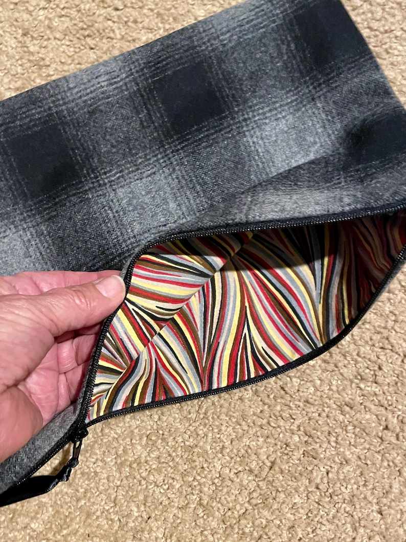 May include: A gray plaid zippered pouch with a black zipper. The interior lining features a colorful striped pattern in red, yellow, gray, and brown. The pouch is open, revealing the lining.