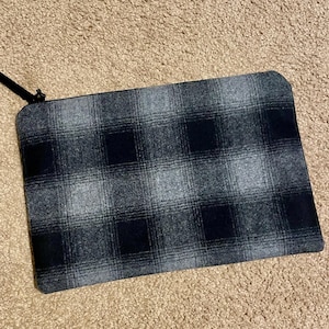 May include: A rectangular, plaid-patterned pouch in shades of gray and black. The pouch has a black zipper and a small black fabric pull. The fabric has a soft, textured appearance. The pouch is likely designed for carrying small items.