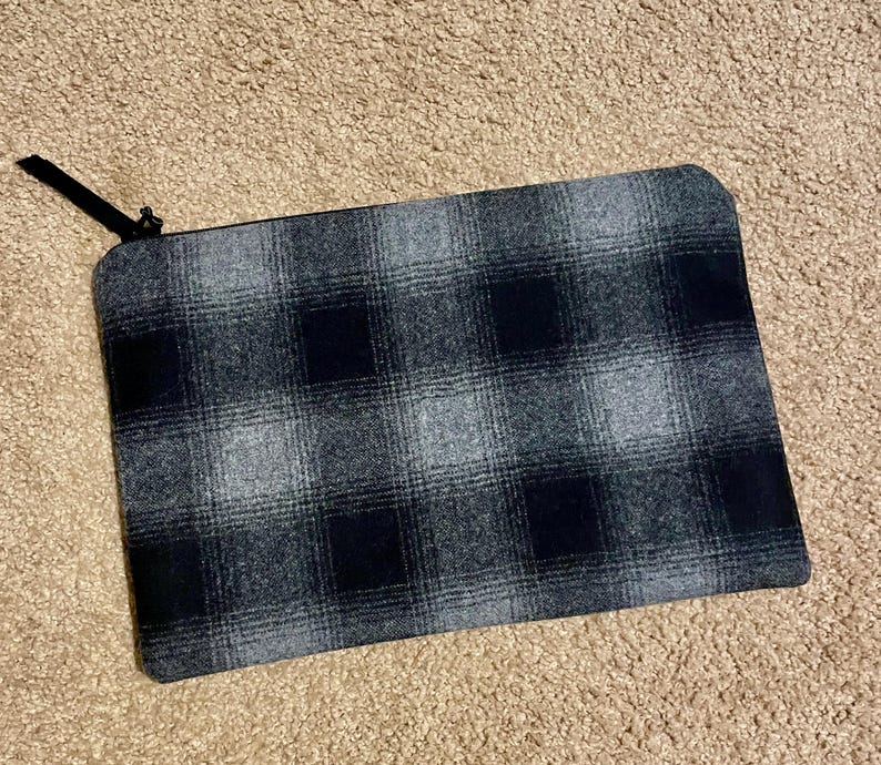 May include: A rectangular, plaid-patterned pouch in shades of gray and black. The pouch has a black zipper and a small black fabric pull. The fabric has a soft, textured appearance. The pouch is likely designed for carrying small items.