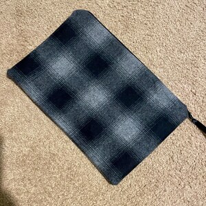 May include: A rectangular, gray and black plaid pattern clutch with a black zipper and a black fabric pull. The clutch is made of a soft, textured fabric. The background is a light brown carpet.