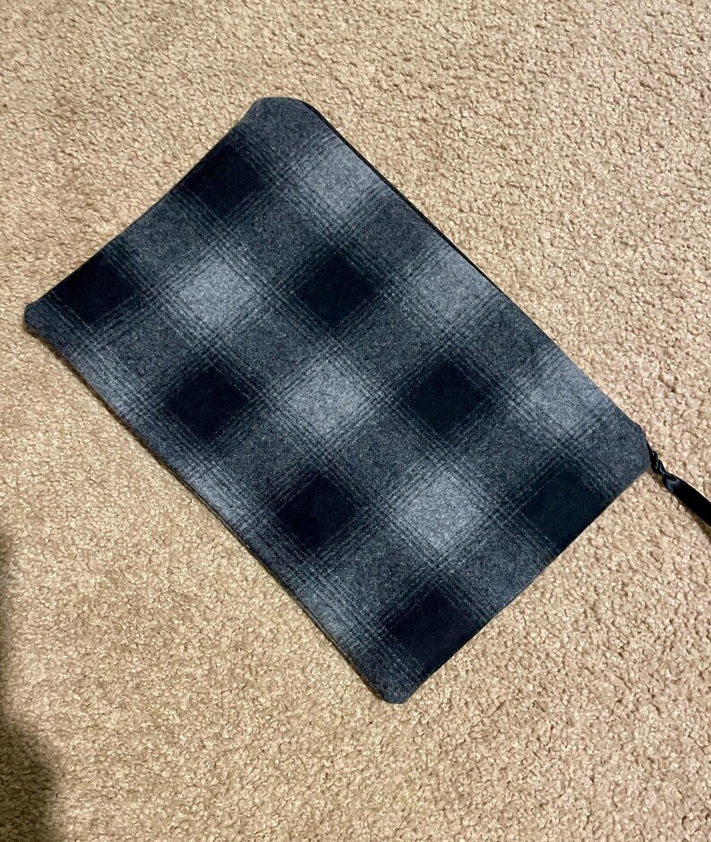 May include: A rectangular, gray and black plaid pattern clutch with a black zipper and a black fabric pull. The clutch is made of a soft, textured fabric. The background is a light brown carpet.