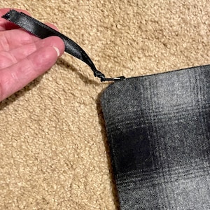 May include: A gray plaid zippered pouch with a black satin ribbon pull. The pouch is made of a woven fabric with a repeating pattern of black and gray squares. The zipper is black.