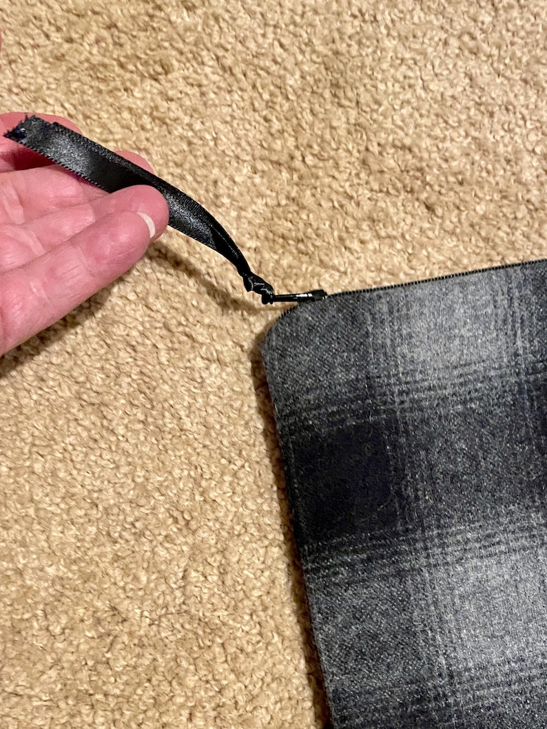May include: A gray plaid zippered pouch with a black satin ribbon pull. The pouch is made of a woven fabric with a repeating pattern of black and gray squares. The zipper is black.