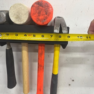 May include: A black metal tool holder with five tools hanging from it. The tools include a hammer with a black handle, a hammer with a wooden handle, a hammer with a red handle, a hammer with a yellow handle, and a claw hammer with a yellow handle. A yellow tape measure is being used to measure the length of the tool holder. The tape measure reads 11 inches.