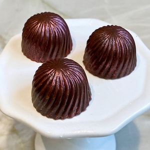 Puede incluir: Tres trufas de chocolate negro con un diseño en espiral y un brillo metálico sobre una fuente blanca escalonada. Las trufas tienen un color marrón rojizo intenso, y la fuente tiene un diseño sencillo y elegante.