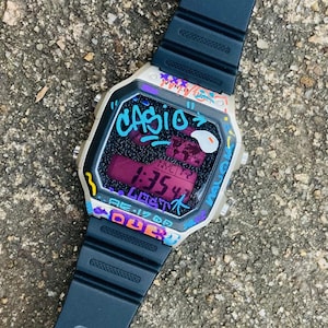 May include: A Casio digital watch with a black strap and a graffiti-style design. The watch face displays the time and city information in a purple hue, with the word "CASIO" in turquoise. The watch is set against a textured grey surface.
