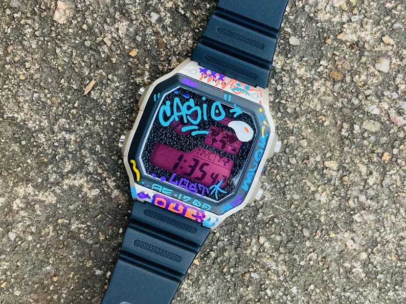 May include: A Casio digital watch with a black strap and a graffiti-style design. The watch face displays the time and city information in a purple hue, with the word "CASIO" in turquoise. The watch is set against a textured grey surface.