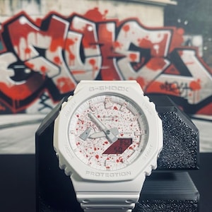 May include: White G-Shock watch with a splatter design on the face, set against a graffiti background. The watch features a digital display and analog hands. The watch is on a black stand.