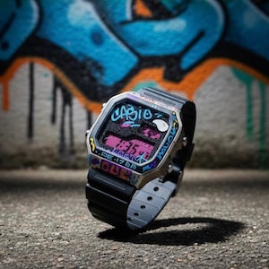 May include: A Casio digital watch with a black strap and a silver-tone case. The watch face displays the time in pink digits and is decorated with graffiti-style art in blue, pink, and yellow. The background features a colourful graffiti mural.