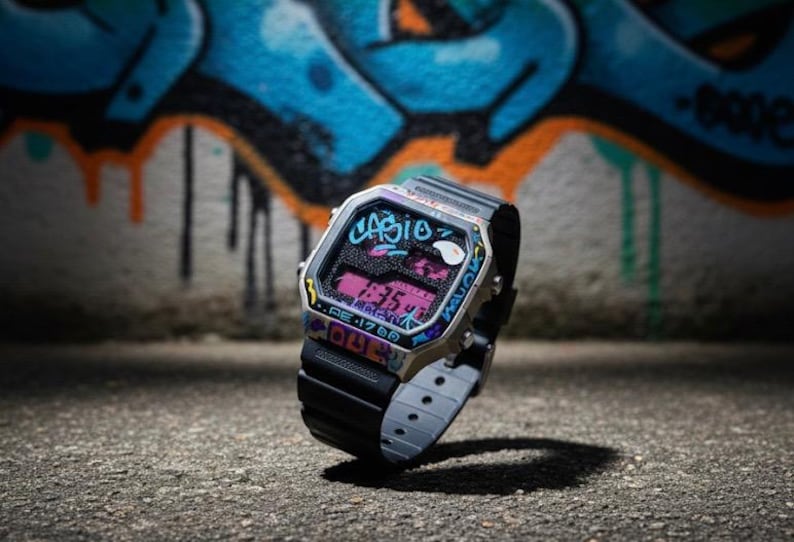May include: A Casio digital watch with a black strap and a silver-tone case. The watch face displays the time in pink digits and is decorated with graffiti-style art in blue, pink, and yellow. The background features a colourful graffiti mural.