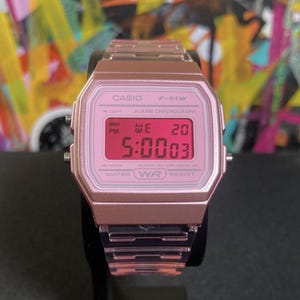 May include: A rose gold Casio F-91W digital watch displaying the time as 5:00:03 PM on Wednesday the 20th. The watch has a rectangular face and a matching metal band. The watch is water resistant.