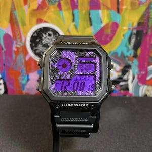 Puede incluir: Reloj digital negro con esfera cuadrada y pantalla morada. El reloj presenta el texto "WORLD TIME" e "ILLUMINATOR". La correa del reloj es negra.