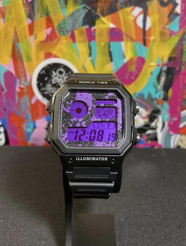 Puede incluir: Reloj digital negro con esfera cuadrada y pantalla morada. El reloj presenta el texto "WORLD TIME" e "ILLUMINATOR". La correa del reloj es negra.