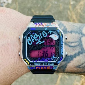 May include: A Casio digital watch with a black strap and a cream-coloured case. The watch face displays the time and is decorated with graffiti-style art in various colours. The watch reads 1:36.