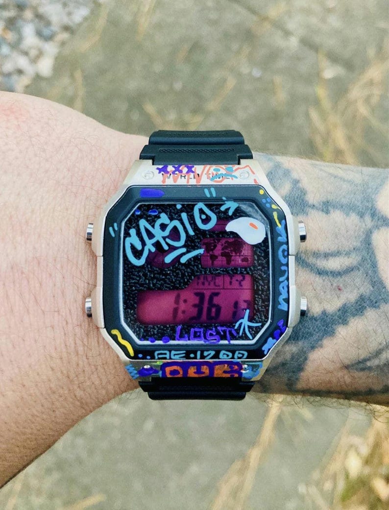May include: A Casio digital watch with a black strap and a cream-coloured case. The watch face displays the time and is decorated with graffiti-style art in various colours. The watch reads 1:36.