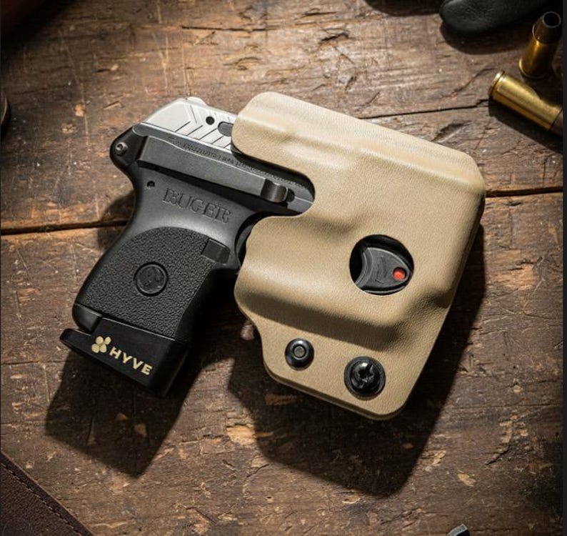 May include: A black Ruger handgun with a silver slide is partially holstered in a tan-colored holster. The magazine base is black with the word "HYVE" printed on it. The image is set on a wooden surface.