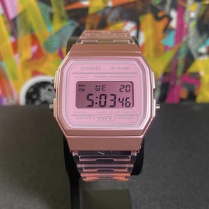 May include: A pink Casio F-91W digital watch. The watch displays the time, day, and date on a digital screen. The watch has a pink resin band and case. The watch is water resistant.
