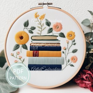May include: A circular wooden embroidery hoop containing white fabric with a hand-stitched design of a stack of six books surrounded by yellow, peach, and green floral elements. The text PDF PATTERN is visible in a light blue circle on the left.