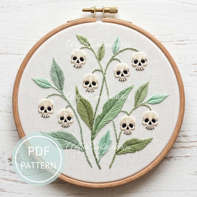 Puede incluir: Un bastidor de bordado circular de madera que contiene tela blanca con un dise&ntilde;o bordado a mano de hojas y tallos verdes con peque&ntilde;as calaveras de color crema como flores. Se observa un gr&aacute;fico circular azul claro con el texto PDF PATTERN.