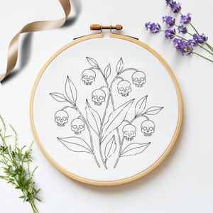 Puede incluir: Un bastidor de bordado circular de madera que contiene tela blanca con un dise&ntilde;o de arte lineal negro de flores de lirio de los valles donde las flores en forma de campana son reemplazadas por peque&ntilde;as calaveras humanas, rodeadas de hojas y tallos verdes.