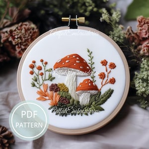 Può includere: Un telaio da ricamo contenente un pannello di tessuto bianco con un disegno ricamato a mano di due funghi arancioni con macchie bianche, circondati da fogliame verde e fiori arancioni. Il testo PDF PATTERN è visibile in un cerchio azzurro.