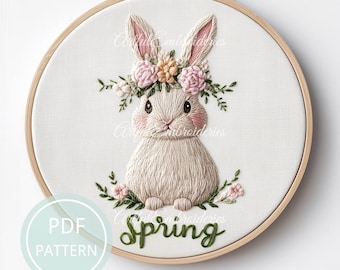 Puede incluir: Arte de bordado en aro con un conejito blanco que lleva una corona floral rosa y verde. El conejito está rodeado de hojas y flores verdes. El texto "Spring" está bordado en verde debajo del conejito.