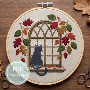 May include: An embroidery hoop containing a linen fabric with a stitched design of a black cat sitting on a windowsill looking out of an arched window. The window is framed by embroidered autumn leaves in shades of red, orange, and green. Text reads PDF PATTERN.