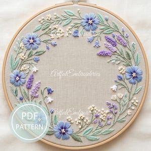 May include: A hand-stitched embroidery hoop featuring a floral wreath design. The wreath includes blue, purple, and white flowers with green leaves, all set against a beige fabric background. The text "Artful Embroidieries" is visible. A PDF pattern is also available.
