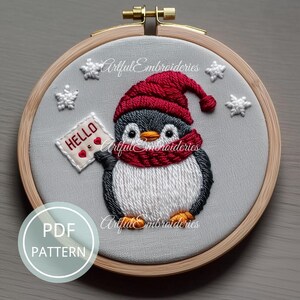 May include: A circular wooden embroidery hoop containing a fabric piece with a hand-stitched penguin wearing a red knit hat and scarf. The penguin holds a white sign with the text HELLO and two red hearts. White stitched snowflakes surround the penguin.