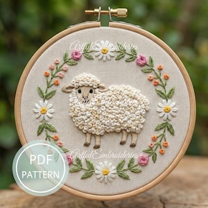 May include: A cream-coloured embroidered sheep in a wooden hoop, encircled by a floral wreath of pink roses, white daisies, and orange buds. The text "Artful Embroidieries" is visible. A PDF pattern is also available. The hoop is approximately 20 cm in diameter.