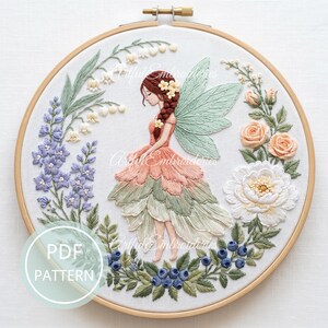 May include: A circular wooden embroidery hoop containing a fabric panel with a hand-stitched design of a fairy with braided hair and green wings, surrounded by embroidered lily of the valley, blue flowers, peach roses, and blueberries.