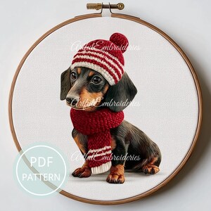 May include: A circular wooden embroidery hoop containing a fabric panel with a stitched illustration of a black and tan dachshund puppy wearing a red and white striped knit hat and a matching red scarf. The text PDF PATTERN is visible.