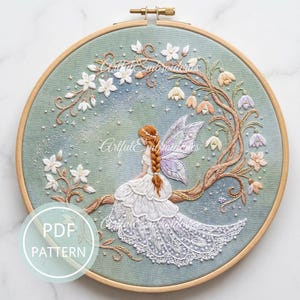 May include: An embroidery hoop containing a fabric piece with a fairy figure sitting on a tree branch. The fairy has auburn hair, a white dress, and purple wings. The scene includes white and pastel flowers, small beads, and the text PDF PATTERN.