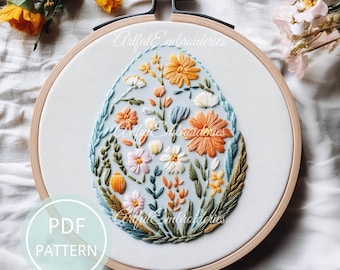 May include: An embroidery hoop containing white fabric with a hand-stitched egg-shaped floral design in shades of orange, yellow, pink, and green. The text ArtfulEmbroidery is visible in the center, and a circular badge reads PDF PATTERN.