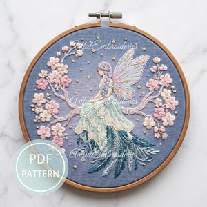 Peut inclure: Un tambour à broder en bois circulaire contenant un panneau en tissu bleu avec un motif brodé à la main représentant une fée aux ailes pastel, assise parmi des branches de cerisier roses et de petites étoiles dorées. Texte PDF PATTERN visible.