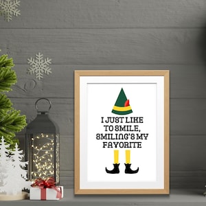 May include: A framed print featuring a green elf illustration and the text "I just like to smile, smiling's my favourite". The print is in a light wood frame and is displayed on a shelf with a small Christmas tree and a gift box.