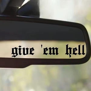 Op de afbeelding: Een zwarte achteruitkijkspiegel met de tekst "give 'em hell" in een gotisch lettertype. De tekst is gecentreerd en duidelijk zichtbaar. De achtergrond is wazig, wat wijst op een auto-interieur.