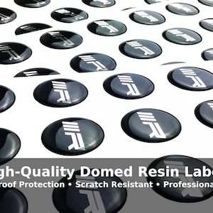 May include: An array of black, oval-shaped domed resin labels with a white graphic. The labels are scattered across a white surface. The text "High-Quality Domed Resin Labels" is visible at the bottom of the image.