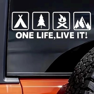 Può includere: Adesivo in vinile bianco con icone a tema campeggio: una tenda, un albero di pino, un falò e montagne. Il testo "ONE LIFE, LIVE IT!" è visualizzato sotto. L'adesivo è applicato a un finestrino del veicolo.