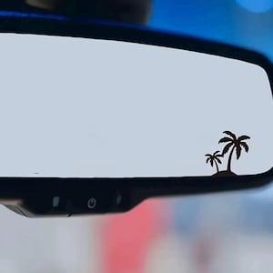Puede incluir: Espejo retrovisor negro con una calcomanía de palmeras. La calcomanía presenta dos palmeras en una pequeña isla. El espejo es rectangular con esquinas redondeadas y tiene botones de control en la parte inferior.