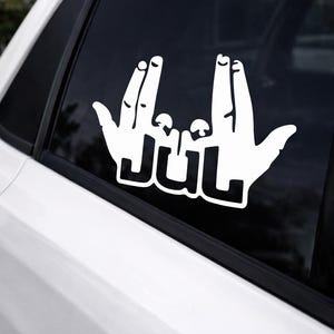 Può includere: Adesivo per auto in vinile bianco con due mani che fanno un gesto con le dita e la parola "JUUL" in grassetto. L'adesivo è progettato per essere applicato su un finestrino dell'auto.