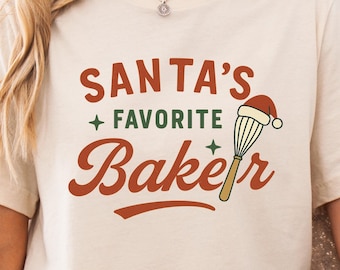 May include: Beige t-shirt with the text "SANTA'S + FAVORITE Baker" in a festive, holiday-themed design. The design includes a whisk wearing a Santa hat. The shirt is a neutral colour and suitable for Christmas baking.