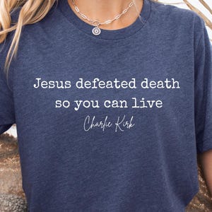 Puede incluir: Camiseta azul marino con el texto blanco "Jesus defeated death so you can live Charlie Kirk". La camiseta tiene cuello redondo y mangas cortas. La persona lleva un collar plateado.