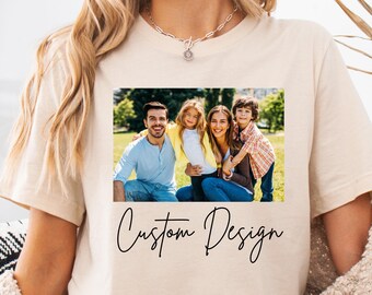 May include: Beige t-shirt featuring a family photo and the words "Custom Design" in a cursive font. The photo shows a family of four smiling. The t-shirt is a neutral color.