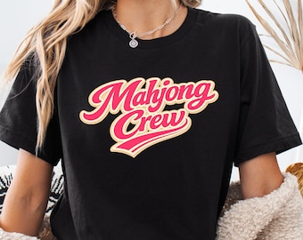 Puede incluir: Camiseta negra con el texto "Mahjong Crew" en una fuente retro y cursiva. El texto es rosa con un contorno amarillo. La camiseta es de estilo casual, adecuada para el uso diario.