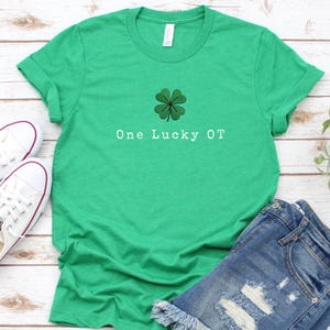以下が含まれることがあります： 「One Lucky OT」の文字と四つ葉のクローバーのグラフィックがプリントされたグリーンのTシャツ。デニムショーツと白いスニーカーとコーディネートされています。Tシャツは柔らかい素材で作られています。