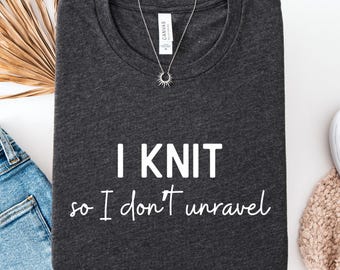 Op de afbeelding: Donkergrijs t-shirt met de witte tekst "I knit so I don't unravel". Een zilveren ketting met een zonnesterhanger ligt op het shirt. Het shirt is op een wit oppervlak gevouwen met een spijkerbroek.